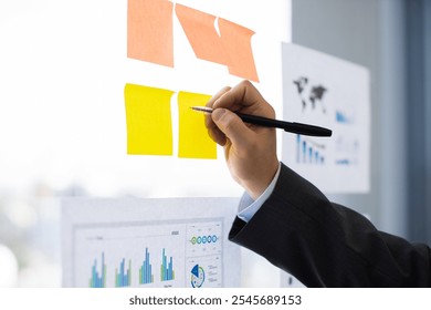 Business professional, possibly male, writing on yellow sticky notes during office meeting. Focused on charts and graphs on transparent board. - Powered by Shutterstock