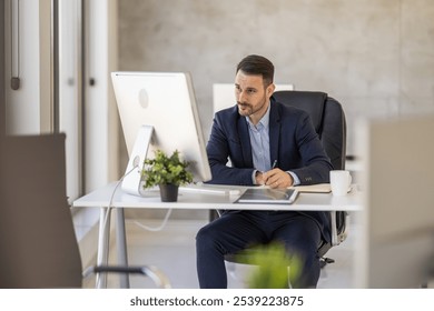 A business professional in a suit focuses intently on his computer screen while working at a modern office desk, with green plant decor adding a fresh touch. - Powered by Shutterstock