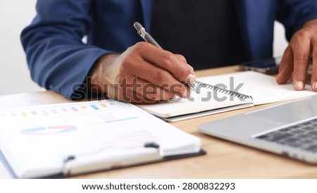 Business professional taking notes while reviewing financial reports at a modern office desk with laptop. Concept of business planning, data analysis, strategy development, productivity, and corporate management.