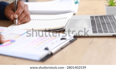 Business professional taking notes while analyzing financial reports and performance charts at office desk, representing data analysis, strategic planning, productivity, reporting, and business management.