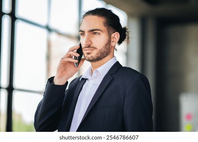 A business professional wearing a suit is talking on a mobile phone inside a modern office setting. Sunlight streams through large windows, creating a dynamic atmosphere. - Powered by Shutterstock