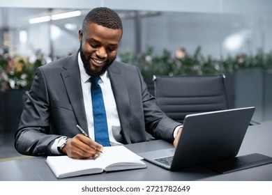 Business Professional Writing Notes While Using a Laptop in a Modern Office Setting