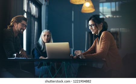 Business Professionals Sitting Around Table And Looking At Laptop. Asian Female Executive Giving A Presentation Over Laptop To Colleagues At Office.