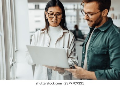 Business professionals standing in an office and using a laptop together. Two young business people having a discussion in a workplace.