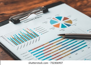 Business report summary on clipboard with colorful bar charts, pie chart, and pen on wooden table. Financial analysis, marketing data, performance review, annual report concept background.