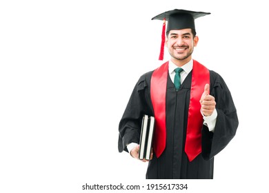 Business School Student Holding Academic Books Under His Arm And Giving A Thumbs Up. Happy Young Latin Man In A Graduation Gown After Receiving His University Diploma