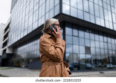 Business Senior Adult Woman In An Autumn Coat On The Background Of The Glass Facade Of The Office Center Speaks On A Mobile Phone, 50 Year Old European Woman, Business Strategy Concept