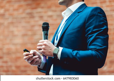 Business and speech topic: Man in a blue suit holding a gray microphone a on a orange bricks background