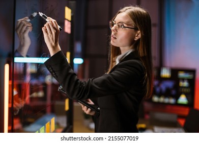 Business, startup, planning and people concept. Pretty businesswoman in eyeglasses drawing on glass board with stickers, working on project tasks at modern night office. Deadline concept - Powered by Shutterstock