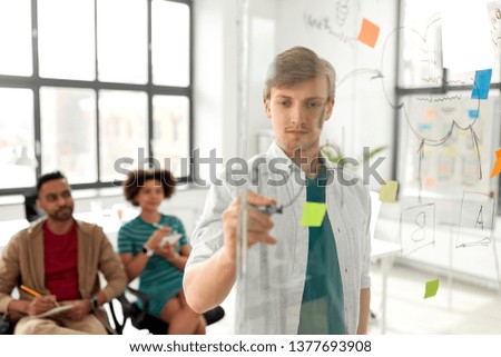 business, startup, planning and people concept - happy man drawing scheme on glass board for creative team at presentation in office