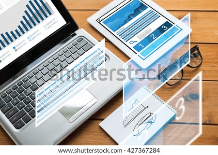 business, statistics and technology concept - close up of on laptop computer, tablet pc, notebook and eyeglasses with charts on wooden table