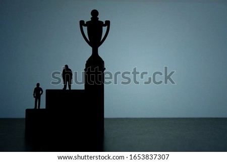 Business strategy conceptual photo - Silhouette of miniature businessman standing on podium to reach winning trophy