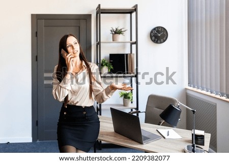 Business talk. Cheerful young beautiful woman talking on mobile phone and looking to the window while leaning on the table at her working place.