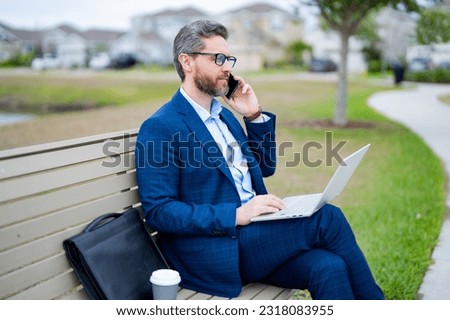 Business talk. Business man talk on phone sitting on a bench in park. Man in suit call phone outside. Handsome business man talk phone sit on park bench. Business man in suit talk on phone in park.