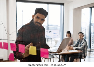 Business team brainstorming session with sticky notes on transparent board. Mid-adult man leading discussion with laptop while colleagues observe from table. Modern office setting. - Powered by Shutterstock