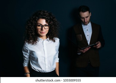 Business Team, Curly Woman With Glasses Leader And Young Male Assistant On A Dark Background.