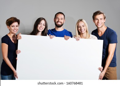 Business Team Or Group Of Diverse Young Professional Friends Holding A Blank White Sign In Front Of Them With Friendly Smiles For Your Text Or Advertisement