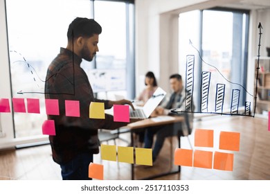 Business team having meeting with charts and post-it notes. Man using laptop with colleagues in background discussing business strategy and data analysis. - Powered by Shutterstock