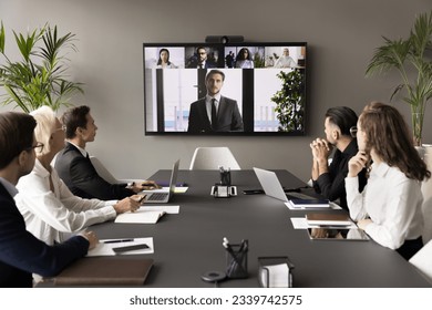 Business team and leader meeting online and offline, sitting at conference table, looking at interactive board with headshots, listening to speaker, CEO, discussing project on group video call - Powered by Shutterstock