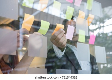 Business team look at the adhesive notes on glass wall in meeting room. Sticky note paper reminder schedule for discussing idea in office. teamwork, brainstorming concept. shot through window