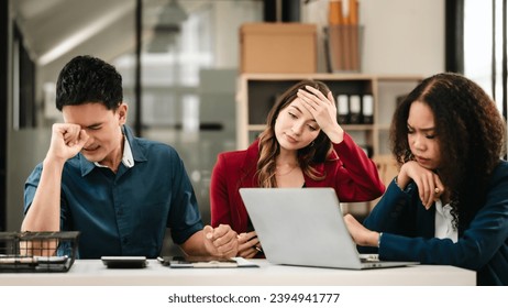 Business team meeting with stressed man rubbing his forehead, while two women review documents, appearing serious and focused. - Powered by Shutterstock