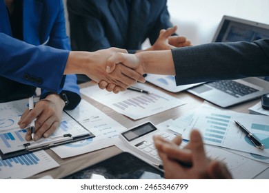 In a business team meeting, success is celebrated with a handshake. Hands close up, dressed in formal suits, working at desks with financial papers, calculators, and laptops. - Powered by Shutterstock