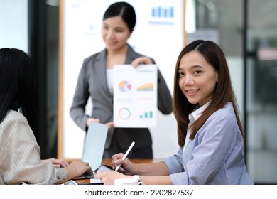  Business. Team Meetings. A Group Sitting Down Around A Table. One Person In The Foreground. A Confident Businesswoman