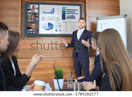 Business team at office, businessman presenting and discussing successful financial report of their company, on screen of plasma TV at meeting room, talking to his colleagues in video conference.