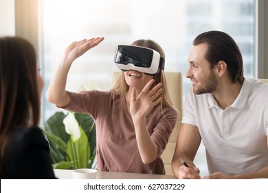 Business team of three people working on virtual reality applications and games, young excited woman testing VR glasses or goggles sitting in the office room with two colleagues, teleconference 