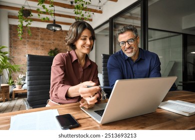 Business team of two executives working together using laptop in office. Happy young business woman manager showing project presentation to mature man client, investor or partner at work meeting.