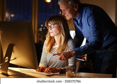 Business Team Working Late At Office. Middle Age Sale Assistant Sitting In Front Of Computer. Senior Businessman Standing Next To Her And And Showing Something With His Finger At Monitor. Teamwork. 