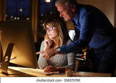 Business Team Working Late At Office. Middle Age Sale Assistant Sitting In Front Of Computer. Senior Businessman Standing Next To Her And And Showing Something With His Finger At Monitor. Teamwork. 