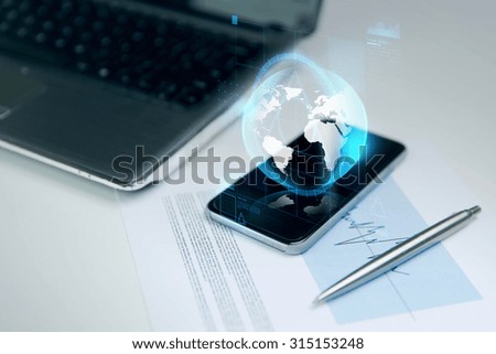 business, technology and communication concept - close up of smartphone with earth globe hologram, laptop computer and chart with pen on office table
