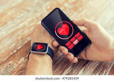 Business, Technology, Health Care And People Concept - Close Up Of Male Hand Holding Smart Phone And Wearing Smart Watch Showing Red Heart Beat Icon On Screen