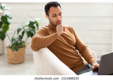 Business And Technology. Portrait Of Focused Middle Eastern Man In Headphones Working On Laptop Computer At Home, Serious Guy Sitting On Couch And Using Pc, Looking At Screen, Watching Webinar