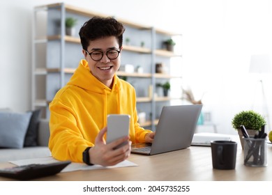 Business And Telecommuting Concept. Portrait of smiling young asian man using smartphone and modern laptop computer, reading text message, sitting at desk with gadgets, working at home office
