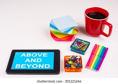 Business Term / Business Phrase On Tablet PC - Colorful Rainbow Colors, Cup, Notepad, Pens, Paper Clips, White Surface - White Word(s) On A Cyan Background - Above And Beyond