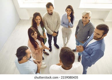 Business Trainer Explaining Something To Group Of People. Team Of Happy Businessmen And Businesswomen Standing In Circle Listening To Coach During Training Meeting. From Above, Overhead, High Angle