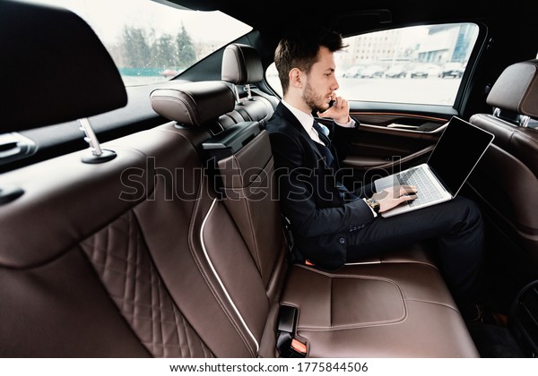 Business Travel. Busy chief\
executive in suit using laptop, making phone call in luxury car,\
side view