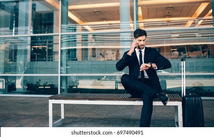 Business Traveler Talking On Mobile Phone And Looking At His Watch.  Businessman Sitting On Bench Making Phone And Checking Time Outside Public Transport Building.