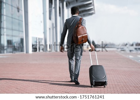 Business Trip Concept. Black Businessman With Travel Suitcase And Backpack Walking Back To Camera Leaving Airport. Copy Space