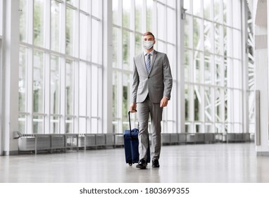 Business Trip, Health And People Concept - Young Businessman Walking With Travel Bag Along Office Building Or Airport Wearing Face Protective Medical Mask For Protection From Virus Disease