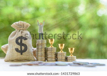 Business tycoon, career success concept : Miniature figurine businessman raises his hands, US dollar bags, golden trophy cups on rows of increasing coins, depicts the ambition obtain intended results