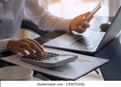 Business woman or accountant hand using calculator, working on mobile smart phone and laptop computer with documentary data graph paper on the table at modern home office. Online marketing concept.