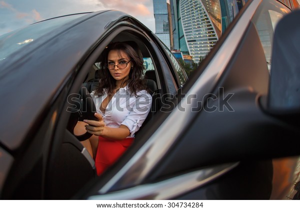 Business woman in a black car on drivers seat after\
hardworking day. A Brunette Lady sitting in a luxury vehicle\
looking from open\
door.