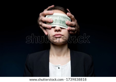 Business woman blinded by money. Dollars close her eyes and hands out of the darkness press money to her face