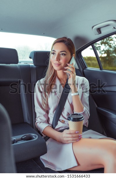 Business woman in car taxi, pink tanned leather\
suit, ringing on cell phone, meeting friends, renting car sharing\
car. Strict guest lady business, cup of coffee tea, trip to an\
important meeting