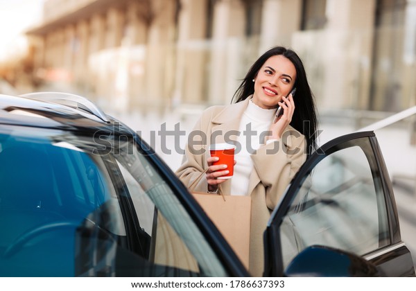 Business woman drinking coffee and talking on the\
phone near her car