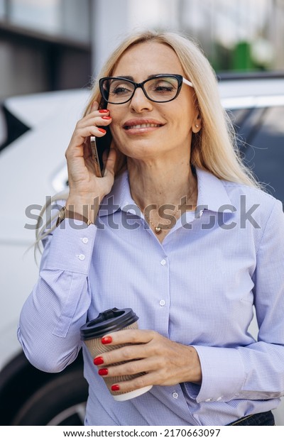 Business woman drinking coffee and talking on\
the phone by the car\
showroom