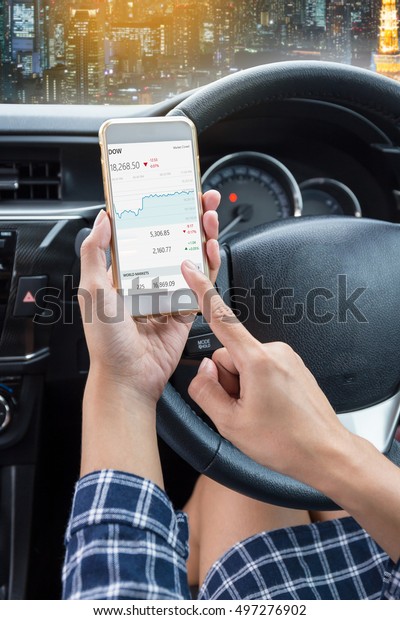 Business woman driver a touch screen of\
white smartphone check stock market or exchange and hand holding\
steering wheel in a car with night city\
background